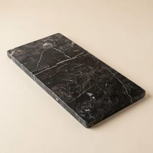May include: A rectangular serving board crafted from dark black marble, featuring prominent white veining throughout the surface. The stone slab has a smooth, polished finish with straight edges and a solid, heavy appearance for food presentation.