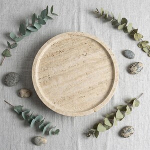 May include: A circular beige travertine tray with a natural porous texture and a raised rim. The stone surface features subtle linear veining and variations in color, showcasing the organic characteristics of the material in a simple round form.