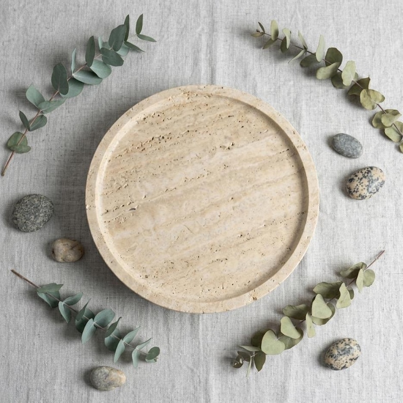May include: A circular beige travertine tray with a natural porous texture and a raised rim. The stone surface features subtle linear veining and variations in color, showcasing the organic characteristics of the material in a simple round form.