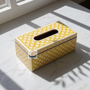 Peut inclure: Une boîte à mouchoirs rectangulaire ornée d'un motif en treillis jaune et blanc sur le dessus et les côtés. La boîte présente une ouverture ovale centrale pour distribuer les mouchoirs et affiche le logo Nestro Exim sur la face avant.