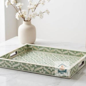 Könnte beinhalten: Ein rechteckiges Serviertablett mit einem geometrischen Blumenmuster in Salbeigrün und Cremeweiß. Das Tablett verfügt über integrierte Seitengriffe und ein flaches Profil. Ein kleines Logo mit dem Text Nestro Exim ist unten rechts zu sehen.