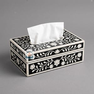 Puede incluir: Una caja de pañuelos rectangular con fondo negro, un patrón de enredadera floral blanca y un borde blanco. Un pañuelo blanco sobresale de la abertura central. La caja muestra un logotipo con el texto Nestro Exim en el centro.