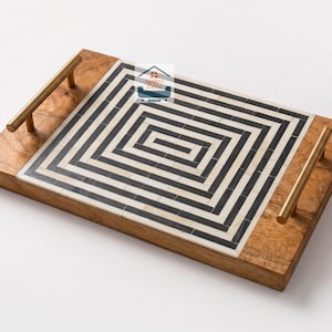 Könnte beinhalten: Ein rechteckiges Serviertablett aus Holz mit einer zentralen Einlage aus schwarz-weißen Fliesen in einem geometrischen Labyrinth-Muster. Das Tablett hat zwei Griffe aus gebürstetem Goldmetall an den Seiten und zeigt den Text Nestro Exim.