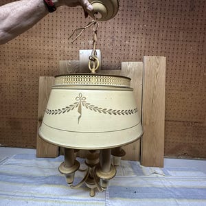 May include: A cream-colored metal pendant light fixture featuring a wide, tapered shade with a decorative gold-toned laurel leaf garland pattern, a perforated top rim, and a central structure with multiple candle-style light sockets.