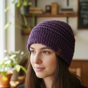 Peut inclure: Un bonnet en tricot violet foncé avec une texture côtelée et une petite étiquette rectangulaire en cuir marron fixée sur le bord inférieur. Le bonnet est fabriqué avec de la laine épaisse et présente une coupe ajustée couvrant la tête.