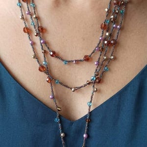 Puede incluir: Un collar de cuentas de múltiples hilos con hilo gris oscuro, intercalado con pequeñas cuentas de color ámbar, turquesa y lavanda. El collar incluye un dije en forma de flor rosa y un colgante circular con un diseño de libélula morada.