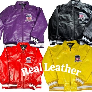 May include: Four leather jackets in purple, black, red, and yellow. Each jacket features a front zipper, a collar, and a patch on the left chest displaying an American flag shield with the text "AVIREX". The text "Real Leather" is visible.