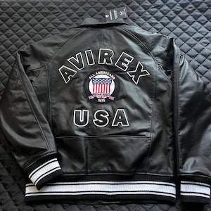 May include: A black leather jacket featuring the text AVIREX and USA in white lettering on the back. A circular patch in the center displays the text ALL AMERICANS ESTABLISHED 1975 with a United States flag. The jacket has black and white striped cuffs.
