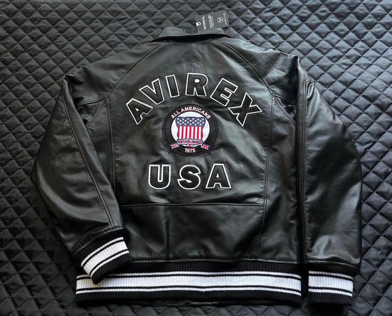 May include: A black leather jacket featuring the text AVIREX and USA in white lettering on the back. A circular patch in the center displays the text ALL AMERICANS ESTABLISHED 1975 with a United States flag. The jacket has black and white striped cuffs.