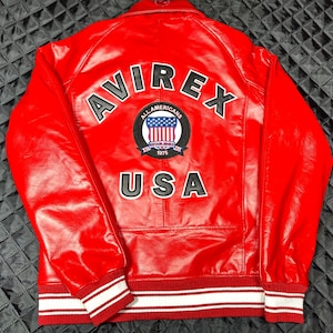 May include: A bright red leather jacket featuring the word AVIREX in large black letters across the upper back. Below this, a circular patch displays the text ALL AMERICANS ESTABLISHED 1975 with a US flag graphic. The word USA appears below the patch.