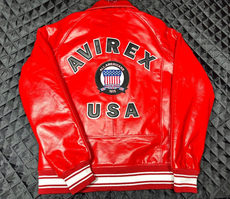 May include: A bright red leather jacket featuring the word AVIREX in large black letters across the upper back. Below this, a circular patch displays the text ALL AMERICANS ESTABLISHED 1975 with a US flag graphic. The word USA appears below the patch.