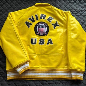 May include: A bright yellow jacket featuring the text AVIREX and USA in black lettering on the back. A circular patch in the center displays the text ALL AMERICANS ESTABLISHED 1975 with a shield graphic. The jacket has white and yellow striped cuffs.