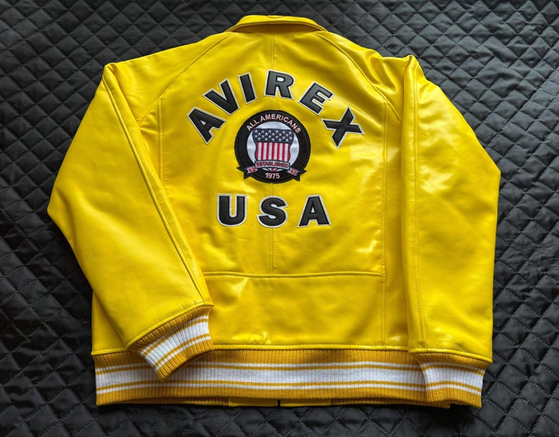 May include: A bright yellow jacket featuring the text AVIREX and USA in black lettering on the back. A circular patch in the center displays the text ALL AMERICANS ESTABLISHED 1975 with a shield graphic. The jacket has white and yellow striped cuffs.