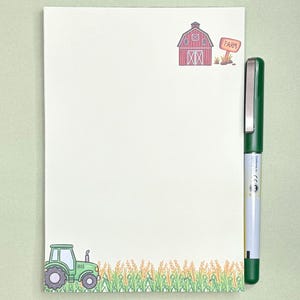 May include: A rectangular notepad featuring a white background with a red barn and a sign labeled FARM in the top right corner, and a green tractor in a field of wheat at the bottom, accompanied by a green pen with a silver clip on the right side.