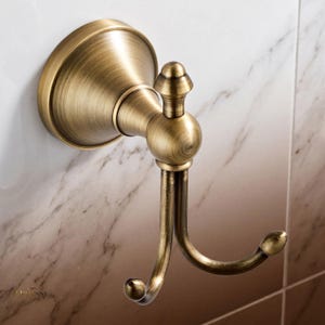 May include: A double-pronged wall hook with a brushed antique brass finish. The hardware features a circular mounting base, a central stem with a decorative finial top, and two curved hooks extending outward, each ending in a rounded tip.