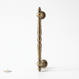May include: A vertical antique brass door handle featuring a slender, elongated central shaft with decorative spherical and tapered accents. The hardware includes two circular mounting plates at the top and bottom. The text ORLIENNE LIVING is visible.