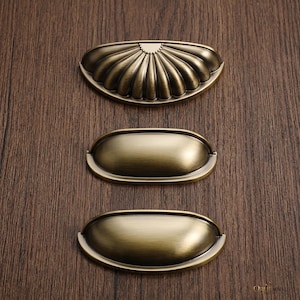 May include: Three antique brass-toned metal drawer pulls arranged vertically. The top pull features a semi-circular shell-like design with radiating ridges. The middle and bottom pulls are smooth, oval-shaped cup handles with a brushed finish.
