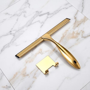 May include: A gold-toned metal squeegee featuring a smooth, ergonomic handle and a black rubber blade, accompanied by a matching gold-toned metal wall-mounted hook with two prongs. The brand name Orlienne Living is printed in the bottom corner.