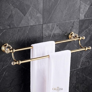 May include: A double-bar towel rack finished in polished gold with clear crystal-style accents at the wall mounts and bar ends. The rack holds two white towels draped over the horizontal bars, featuring a metallic structure and decorative details.