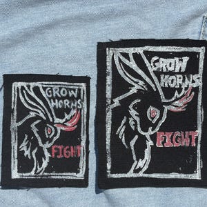May include: Two rectangular black fabric patches featuring a white screen-printed graphic of a horned rabbit. The text GROW HORNS is printed in white above the rabbit, and the word FIGHT is printed in red below. The patches are placed on light blue denim.