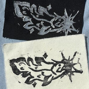 Op de afbeelding: Twee rechthoekige stoffen patches met een gestileerde grafische print van een vis met een sterrenontwerp. De ene patch is zwart met een zilverkleurige inktprint, de andere is naturelkleurig met een zwarte inktprint.