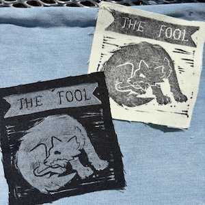 May include: Two square fabric patches featuring a block print design of a curled-up fox. One patch is black with a white print, and the other is off-white with a black print. Both patches display a banner at the top with the text THE FOOL.