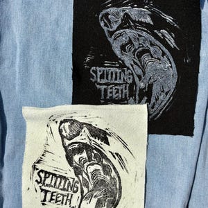 May include: Two square fabric patches featuring a graphic illustration of a snarling mouth with sharp teeth and the text "SPITTING TEETH". One patch is black with white ink, and the other is off-white with black ink, both attached to light blue denim.
