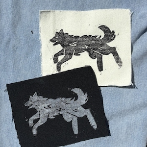May include: Two rectangular fabric patches featuring a screen-printed graphic of a running wolf. One patch is off-white with a black wolf print, and the other is black with a light gray wolf print. Both patches have raw, frayed edges.