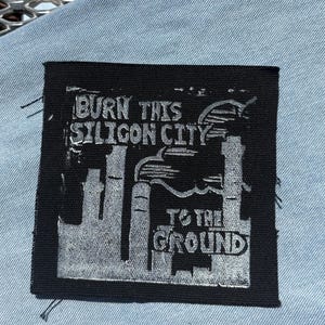 Pode incluir: Um emblema quadrado de tecido preto com um gráfico serigrafado em prata de um horizonte urbano com chaminés. O texto "BURN THIS SILICON CITY TO THE GROUND" está em uma fonte negrito e desgastada, com bordas visivelmente desfiadas no emblema.