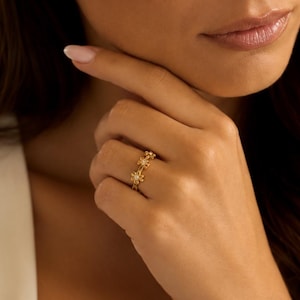 Könnte beinhalten: Ein goldener Ring mit einer Reihe kleiner, miteinander verbundener blumenförmiger Motive. Jede Blume hat einen zentralen, klaren, rund geschliffenen Stein in der Mitte der Blütenblätter. Das Band ist dünn, metallisch und poliert.