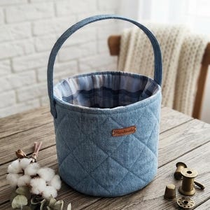 Puede incluir: Una cesta cilíndrica de mezclilla con un patrón de diamantes acolchado y una sola asa. El interior está forrado con una tela de cuadros azules y blancos. Una pequeña etiqueta de cuero marrón con la palabra Handmade está en el frente.