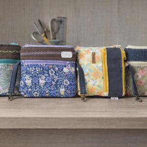 May include: Four small rectangular crossbody bags lined up. The first has a tropical leaf print, the second a blue floral pattern, the third a yellow floral design with a dark blue vertical stripe, and the fourth a soft pink floral print with denim.