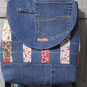 May include: A blue denim backpack featuring a flap with an integrated pocket and a small brown leather tag labeled Handmade. The body is constructed from denim panels interspersed with vertical strips of fabric featuring colorful floral patterns.