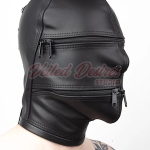 Könnte beinhalten: Eine schwarze Lederhaube mit zwei horizontalen Reißverschlüssen im Gesichtsbereich, einer auf Augenhöhe und einer auf Mundhöhe. Die Haube hat sichtbare Nähte entlang der Kanten und eine glatte Oberfläche, mit dem Text Veiled Desires STUDIO.