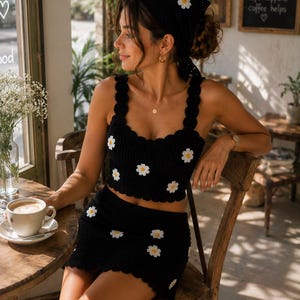 May include: A black crochet two-piece set consisting of a sleeveless crop top and a matching mini skirt, both featuring a scalloped edge and white daisy flower motifs. The set includes a coordinating black crochet headscarf with white daisies.