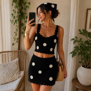 May include: A black crochet two-piece set consisting of a sleeveless crop top and a mini skirt, both featuring a scalloped hem and white daisy flower motifs with yellow centers. A matching black crochet bandana with a white daisy is also included.