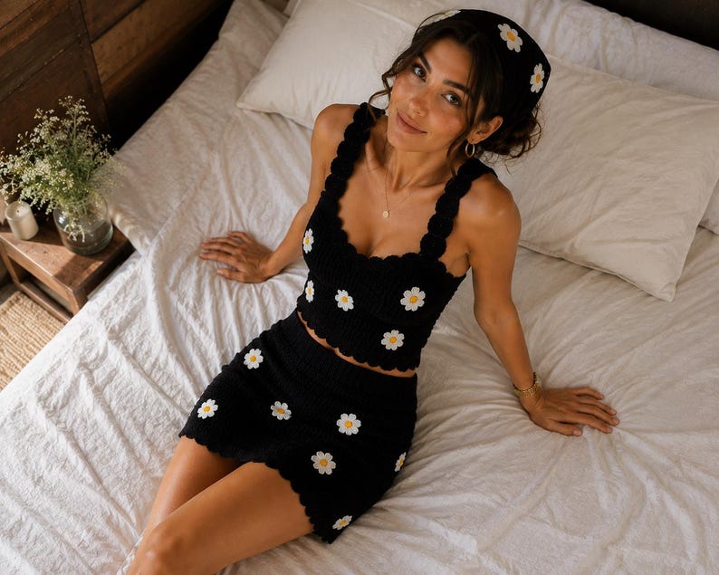 May include: A black two-piece set consisting of a sleeveless crop top and a mini skirt, both featuring a scalloped edge and a repeating white daisy pattern with yellow centers. A matching black headscarf with white daisy accents is also included.