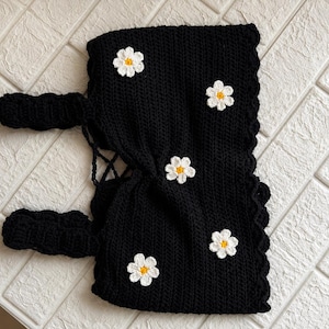 May include: A black crochet top featuring a scalloped edge and four white daisy appliques with yellow centers. The garment includes two thick black crochet straps and a lace-up back closure with thin black cord detailing for an adjustable fit.