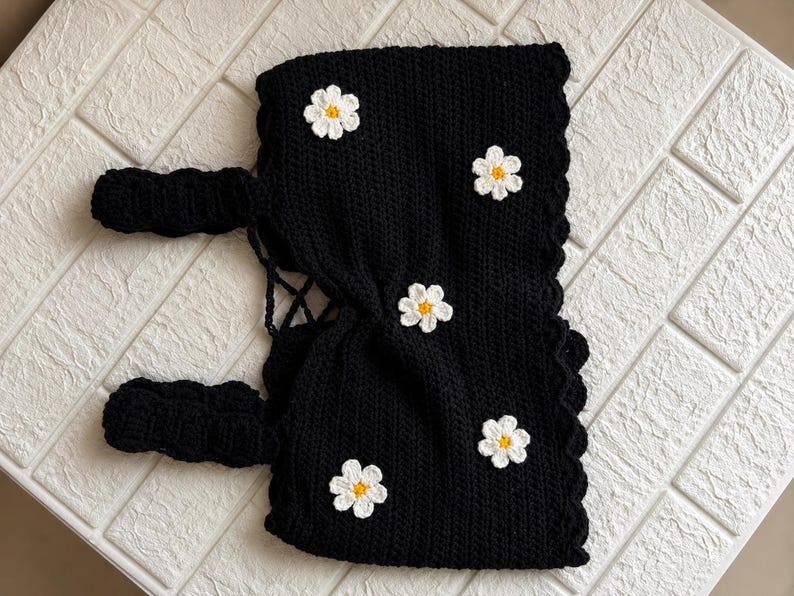 May include: A black crochet top featuring a scalloped edge and four white daisy appliques with yellow centers. The garment includes two thick black crochet straps and a lace-up back closure with thin black cord detailing for an adjustable fit.