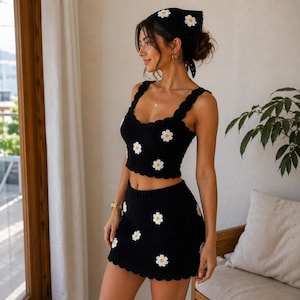 May include: A black crochet two-piece set consisting of a sleeveless crop top and a mini skirt, both featuring a scalloped hem and a repeating pattern of white daisy appliques with yellow centers. A matching black crochet headscarf is also included.