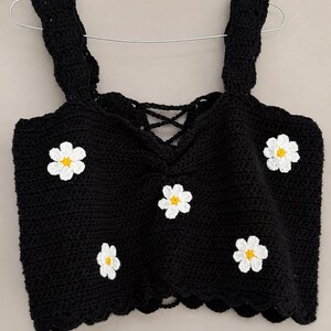 May include: A black crochet crop top featuring a square neckline with a lace-up detail and thick straps. The garment is decorated with five white daisy appliques, each with a yellow center, distributed across the front. The hem has a scalloped edge.