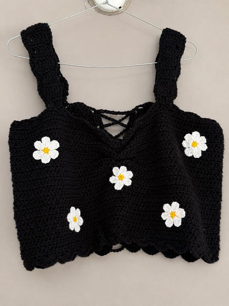 May include: A black crochet crop top featuring a square neckline with a lace-up detail and thick straps. The garment is decorated with five white daisy appliques, each with a yellow center, distributed across the front. The hem has a scalloped edge.