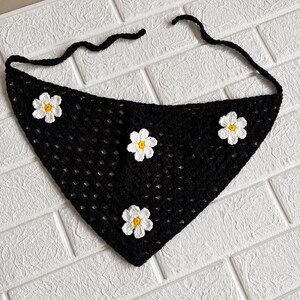 May include: A triangular crochet bandana in black yarn featuring an open-work stitch pattern. The accessory is decorated with four white crocheted flowers, each with a small yellow center. Two long black yarn ties extend from the top corners.