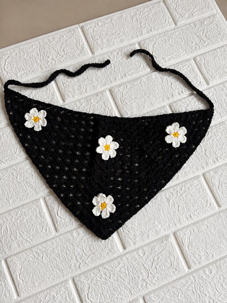 May include: A triangular crochet bandana in black yarn featuring an open-work stitch pattern. The accessory is decorated with four white crocheted flowers, each with a small yellow center. Two long black yarn ties extend from the top corners.