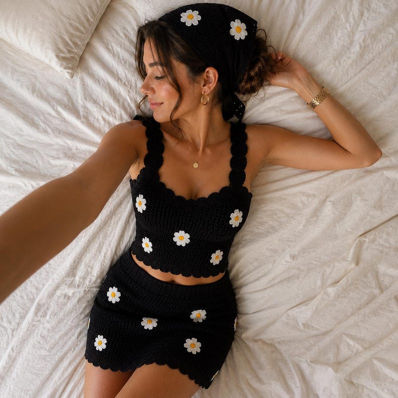 May include: A black crochet two-piece set consisting of a sleeveless crop top and a matching mini skirt. Both pieces feature a scalloped edge and are decorated with white daisy motifs with yellow centers. A matching black crochet headscarf is included.