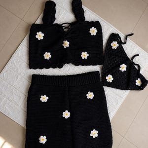 May include: A black crochet set consisting of a sleeveless crop top with thick straps and a lace-up front, a matching skirt with an elasticized waistband, and a triangular headscarf, all decorated with white and yellow daisy flower appliques.