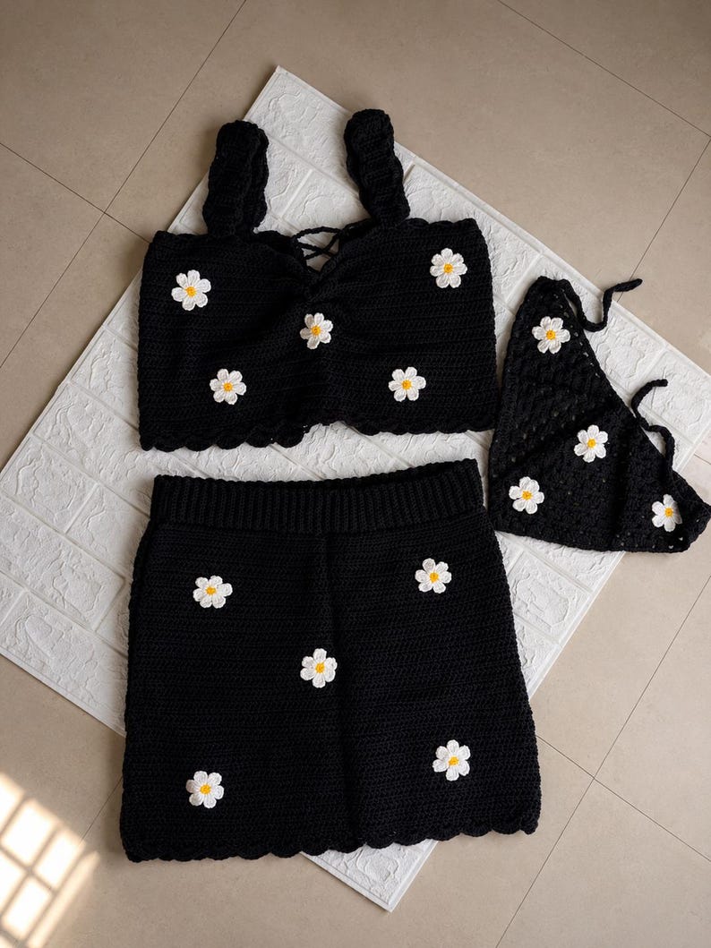 May include: A black crochet set consisting of a sleeveless crop top with thick straps and a lace-up front, a matching skirt with an elasticized waistband, and a triangular headscarf, all decorated with white and yellow daisy flower appliques.