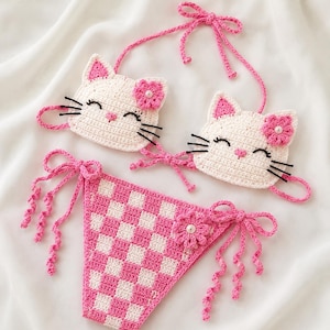 May include: A two-piece crochet bikini set featuring a top with two white cat faces, each with pink inner ears, black embroidered eyes, whiskers, and a pink flower accent. The bottom is a pink and white checkered pattern with side ties.