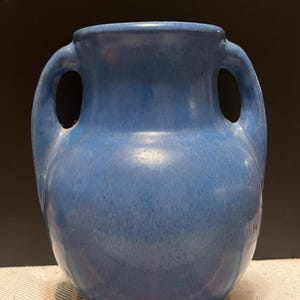May include: A medium-sized ceramic vase with a rounded body and a wide neck. The vase features a solid blue glaze with subtle variations in tone and two symmetrical handles attached to the sides, extending from the neck to the shoulder.