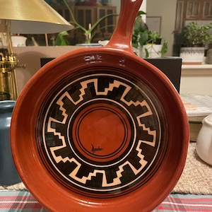 May include: A round, terracotta-colored ceramic skillet with a single handle featuring a hole for hanging. The interior surface displays a glossy finish with a black and cream geometric pattern surrounding a central circle and a signature.